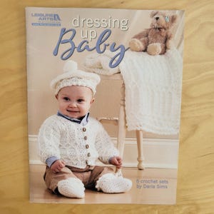 May include: A softcover instructional book titled Dressing Up Baby by Leisure Arts, featuring 5 crochet sets by Darla Sims. The cover displays a baby wearing a white crocheted beret, cardigan, and booties, with a teddy bear and blanket nearby.