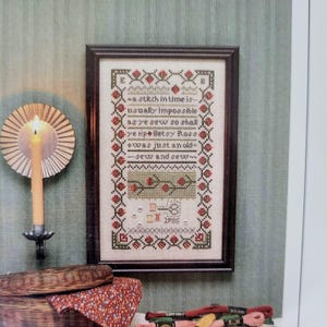 May include: Framed cross-stitch sampler with a dark wooden frame. The sampler features the text "a stitch in time is..." and floral border. A candle and basket are in the foreground. The year "1985" is stitched at the bottom.