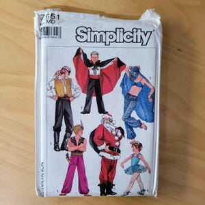May include: A Simplicity sewing pattern packet featuring illustrations of various costume designs. The costumes include a pirate, a vampire with a red cape, a belly dancer, Santa Claus, a genie, and a ballerina. The packet is white with the Simplicity logo.