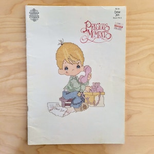 May include: A cross-stitch pattern booklet featuring a Precious Moments character, a young boy, on the phone. The booklet is titled "Dear Jon" and is part of the Precious Moments collection.