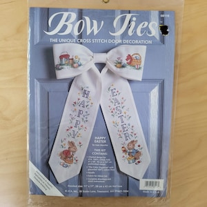 May include: A cross-stitch door decoration kit in a plastic package. The white bow tie features the text "Bow Ties" and "Happy Easter." The design includes Easter bunnies, baskets, and floral accents. Finished size: 11" x 17".