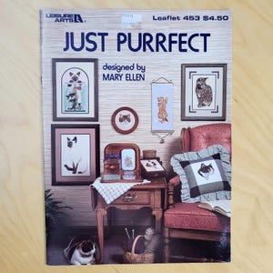 May include: A leaflet titled "JUST PURRFECT" designed by Mary Ellen, featuring cat-themed cross-stitch patterns. The cover displays framed cat portraits, a decorative pillow, and a small table with a basket of yarn.