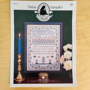 May include: A framed cross-stitch sampler titled "Virtue Sampler" with an alphabet, numbers, and a poem. The design includes floral and tree motifs, with a blue candle and gold candlestick on the left. A small decorative box and flowers are also present.