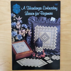 May include: A Designs by Gloria & Pat instructional leaflet titled A Hardanger Embroidery Lesson for Beginners. The cover features a dark blue background with images of Hardanger embroidery projects, including a pillow, a wooden box, and a candle.