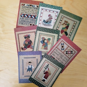May include: A set of eight cross-stitch patterns featuring various designs, including a teddy bear, a Christmas tree, a windmill, and a stocking. The patterns are printed on colorful cardstock with a white border.