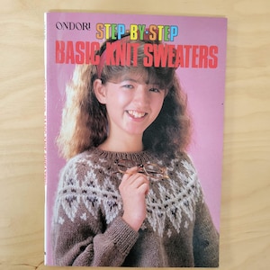 May include: A book cover with the title "STEP-BY-STEP BASIC KNIT SWEATERS" in red and yellow text. The cover features a smiling person wearing a brown sweater with a white and brown geometric pattern, holding glasses.