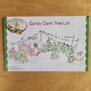 May include: A white paper with a green scalloped border features a colorful embroidered design. The design includes a candy cane, Christmas trees, a snowman, and the text "Candy Cane Tree Lot". The Crab-apple Hill logo is in the upper left corner.