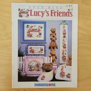 May include: A craft book titled "Lucy's Friends" featuring cross-stitch patterns. The cover displays various finished projects, including framed teddy bear designs, a height chart, and a bib. The color palette includes blues, pinks, and browns, with the Dimensions logo at the bottom.