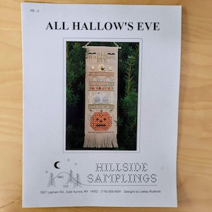 May include: A printed paper with the text "ALL HALLOW'S EVE" at the top. The image features a cross-stitch design with a pumpkin, bats, and the words "ALL HALLOWS EVE". The bottom of the paper has the text "HILLSIDE SAMPLINGS".