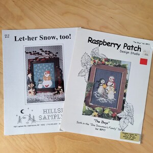 May include: Two cross-stitch patterns featuring winter scenes. The first pattern is titled "Let-her Snow, too!" and depicts a snowman with an alphabet pattern on its belly. The second pattern is titled "The Boys" and is part of the "One Snowman's Family" series. It depicts three snowmen standing together.