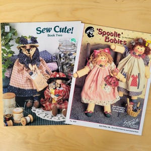 May include: Two craft pattern books titled "Sew Cute! Book Two" and "'Spoolie' Babies" featuring illustrations of handmade dolls and bears. The "Sew Cute!" book shows a bear doll with a shopping bag. The "'Spoolie' Babies" book shows two dolls.
