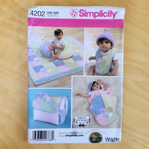 May include: Simplicity sewing pattern 4202 for baby items. The pattern includes instructions for a quilt, bib, hat, and carrier. The pattern features a baby wearing the items. The pattern is in multiple languages.