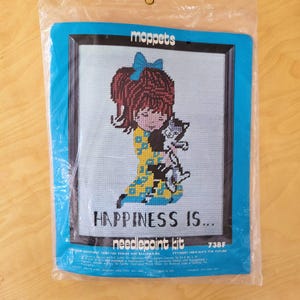 May include: A vintage "moppets" needlepoint kit featuring a girl holding a cat. The design includes a girl with red hair and a blue bow, wearing a yellow dress. The text "HAPPINESS IS..." is below the image.
