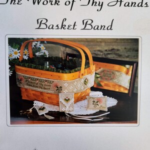 May include: An orange woven basket with a wooden handle, featuring a decorative band with floral embroidery and the text "The work of thy hands comes from thy heart." A framed picture and small embroidered items are also present.