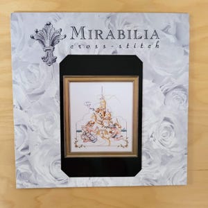 May include: A Mirabilia cross-stitch pattern featuring a framed image of a whimsical scene with figures and a castle. The frame is set against a background of white roses. The text "Mirabilia cross-stitch" is visible.