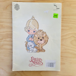 May include: A cross-stitch pattern booklet featuring a Precious Moments illustration of a little girl with blonde hair and a blue dress holding a white flower, standing on a lion. The booklet is titled "Peace on Earth" and is part of the Precious Moments collection.