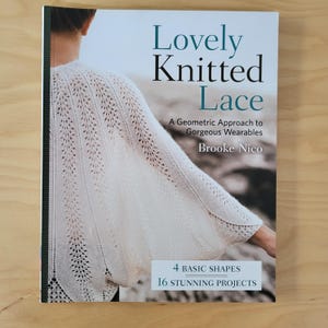May include: A softcover book titled Lovely Knitted Lace: A Geometric Approach to Gorgeous Wearables by Brooke Nico. The cover features a white knitted lace garment with a delicate pattern and includes the text 4 Basic Shapes 16 Stunning Projects.