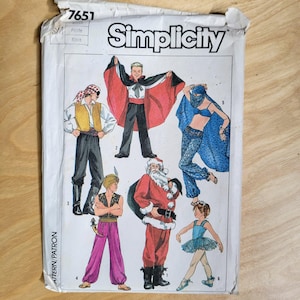 May include: A vintage Simplicity sewing pattern envelope featuring illustrations of various costumes. The costumes include a pirate, a magician with a red and black cape, a Santa Claus, a dancer, and an Arabian Nights-themed outfit. The envelope is white with the word "Simplicity" in large letters.