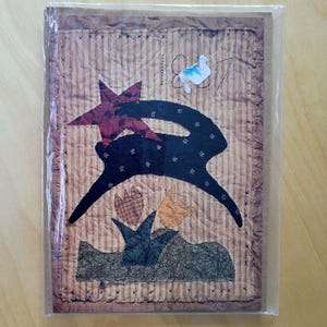 May include: A quilted artwork featuring a black silhouette of a rabbit with star and floral accents. The rabbit is set against a striped background with a red star and floral accents. The artwork is encased in a clear plastic sleeve.