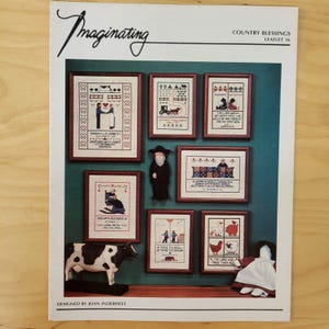 May include: A page from "Imaginating" featuring framed cross-stitch samplers with country themes. The samplers depict couples, farm scenes, and Bible verses. A cow figurine and a doll are also present.
