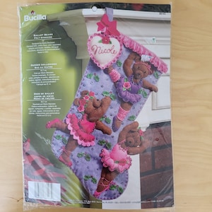 May include: A Bucilla felt stocking kit featuring ballet bears. The stocking is purple with felt bear figures in pink tutus and ballet slippers. A pink heart with the name "Nicole" is attached to the top.