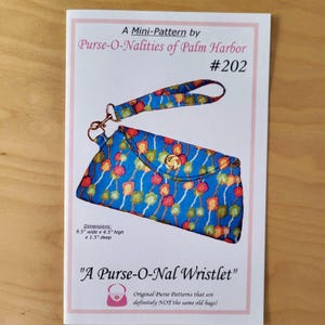 May include: A mini-pattern by Purse-O-Nalities of Palm Harbor for a wristlet purse. The purse is blue with a colorful floral pattern and gold hardware. The dimensions are 9.5 inches wide, 4.5 inches high, and 1.5 inches deep. The text reads "A Purse-O-Nal Wristlet".