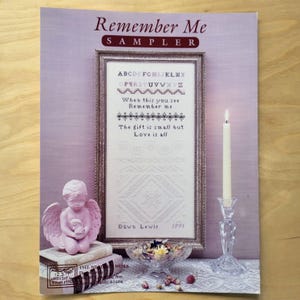 May include: A cross-stitch sampler titled "Remember Me" with a framed design, featuring alphabet letters and a quote. A pink cherub statue, a lit candle in a crystal holder, and a glass bowl with flowers are also present.