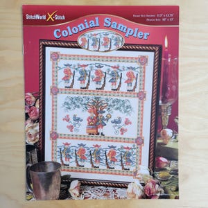 May include: A cross-stitch pattern titled "Colonial Sampler" with a framed design. The design features figures in colonial attire, trees, and birds. The frame size is 11.5 x 13.75 inches, and the design size is 10 x 13 inches. The StitchWorld X-Stitch logo is visible.