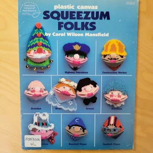 May include: A book cover featuring plastic canvas "Squeezum Folks" patterns by Carol Wilson Mansfield. The cover displays various finished projects, including a clown, highway patrolman, construction worker, bride, and baseball player. The color palette includes blue, green, pink, and yellow.