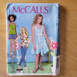 May include: A sewing pattern for girls' dresses and tops. The pattern includes three different styles: a sleeveless dress with a tie waist, a short-sleeved top with a star print, and a sleeveless top with a green bow. The pattern is from McCall's and the pattern number is M7347.