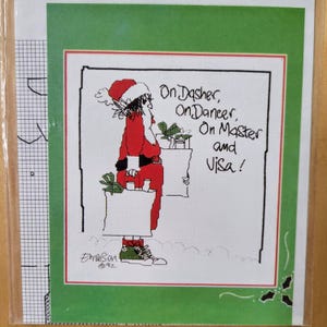 May include: A Christmas-themed cross-stitch pattern featuring a Santa Claus figure in a red suit and hat, carrying a shopping bag. The text reads "On Dasher, On Dancer, On Master and Visa!" The design is framed in a green border.