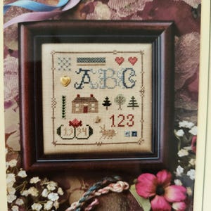 May include: Framed cross-stitch sampler with a dark wood frame. The sampler features the letters ABC, the numbers 123, a house, trees, hearts, and other decorative elements. The background is a light beige fabric.