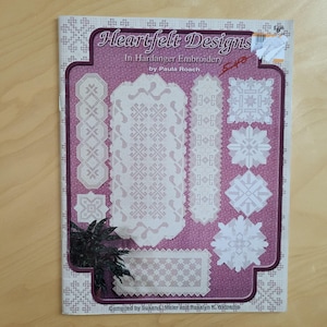 May include: A book titled "Heartfelt Designs" featuring Hardanger embroidery patterns. The cover has a purple background with white embroidered designs in various shapes. The book is by Paula Roach and compiled by Susan E. Meier and Rosalyn K. Watnemo.