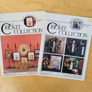 May include: Two cross-stitch pattern books, "The Cricket Collection," are displayed. One is titled "Candlescreens II" and the other "Trimmings Two." The covers feature cross-stitch designs of holiday themes, including framed pictures, wreaths, and a haunted house.