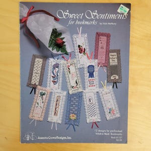 May include: A book cover titled "Sweet Sentiments for bookmarks" by Vicki McMurry. The cover displays various embroidered bookmarks with lace edges and ribbons. A bouquet of flowers is tied with a red ribbon. The book is from Jeanette Crews Designs, Inc.