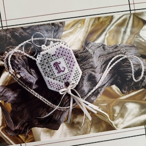 May include: A white fabric scissor fob featuring a cross-stitched heart pattern in shades of purple and green with a central letter L. The fob includes a braided cord, a small dark bead, and white ribbon accents, holding a pair of metal scissors.