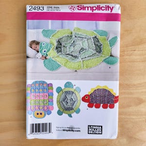 May include: A Simplicity sewing pattern envelope 2493 featuring instructions for three plush animal designs. The cover displays a green turtle-shaped rug, a multi-colored rectangular creature, a circular turtle, and a red-bodied dinosaur-like figure.