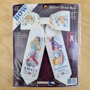 May include: A counted cross-stitch kit for a front door bow. The kit features a cream-colored bow with embroidered angels and the words "Angels Welcome Here." The bow is 17 inches (43 cm) long and includes instructions.