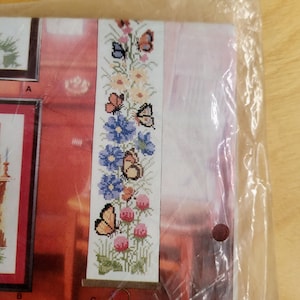 May include: A cross-stitch bookmark featuring a floral and butterfly design. The bookmark has a white background with blue, orange, and pink flowers, and several butterflies. The bookmark is in a plastic package.