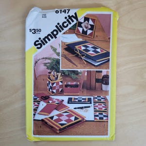 May include: Simplicity sewing pattern envelope with images of quilted accessories, including a pen holder, coasters, and a notebook. The envelope is white with yellow accents and features the text "Simplicity" and "6147".