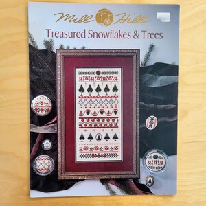 May include: A craft kit titled "Treasured Snowflakes & Trees" featuring a framed cross-stitch design with red, black, and white patterns. The kit includes decorative pins and ornaments with similar designs. The color palette is primarily red, black, and white.