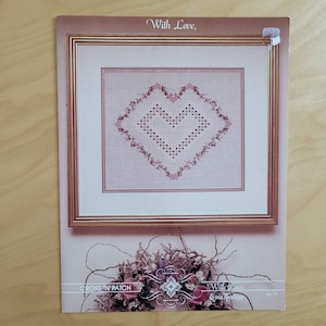 May include: A framed cross-stitch pattern featuring a heart design in shades of pink and white. The words "With Love" are at the top, and the words "Cross 'N Patch" are at the bottom. The pattern is on a light pink background.