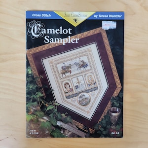 May include: A Just CrossStitch pattern booklet titled Camelot Sampler by Teresa Wentzler. The cover features a finished cross stitch design with knights, a sword, and a castle, framed in dark burgundy and gold, surrounded by green ivy leaves.