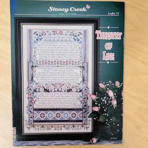 May include: A cross-stitch pattern leaflet titled "Tapestry of Life" by Stoney Creek Collection. The design features floral and geometric patterns in shades of blue, pink, and white, framed in black. The leaflet includes text and is accompanied by a bouquet of pink roses.