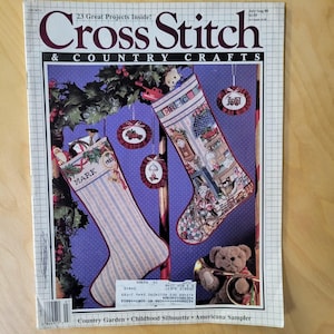 May include: A vintage issue of "Cross Stitch & Country Crafts" magazine. The cover features cross-stitched Christmas stockings with the names "MARK" and "ERIC" on them. The magazine has a purple background with decorative elements and the text "23 Great Projects Inside!"