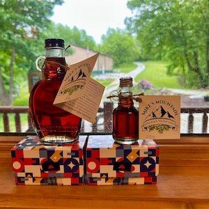 May include: Two glass bottles of Mike's Mountain Pure Black Walnut Syrup, one large and one small, each with a handle and a tied tag. The bottles sit on two matching boxes featuring a geometric pattern of red, blue, yellow, and white shapes.