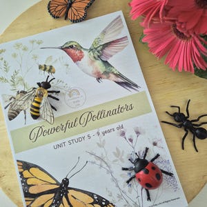 May include: A children's educational unit study titled "Powerful Pollinators" with illustrations of a hummingbird, bees, and butterflies. The cover features the text "UNIT STUDY 5 - 9 years old". Decorative insect figurines and pink flowers surround the book.