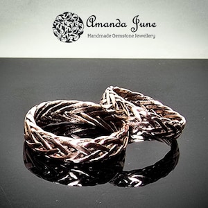 May include: Two bronze-coloured rings with a woven design. The rings are displayed on a reflective black surface. The rings are handmade gemstone jewellery, as indicated by the text "Amanda June Handmade Gemstone Jewellery" in the background.