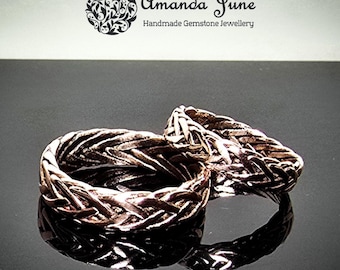May include: Two bronze-colored rings with a woven design. The rings are displayed on a reflective black surface. The rings are handmade gemstone jewelry, as indicated by the text "Amanda June Handmade Gemstone Jewellery" in the background.