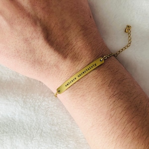 May include: A gold bracelet with a thin chain and a rectangular charm that reads "embrace uncertainty".